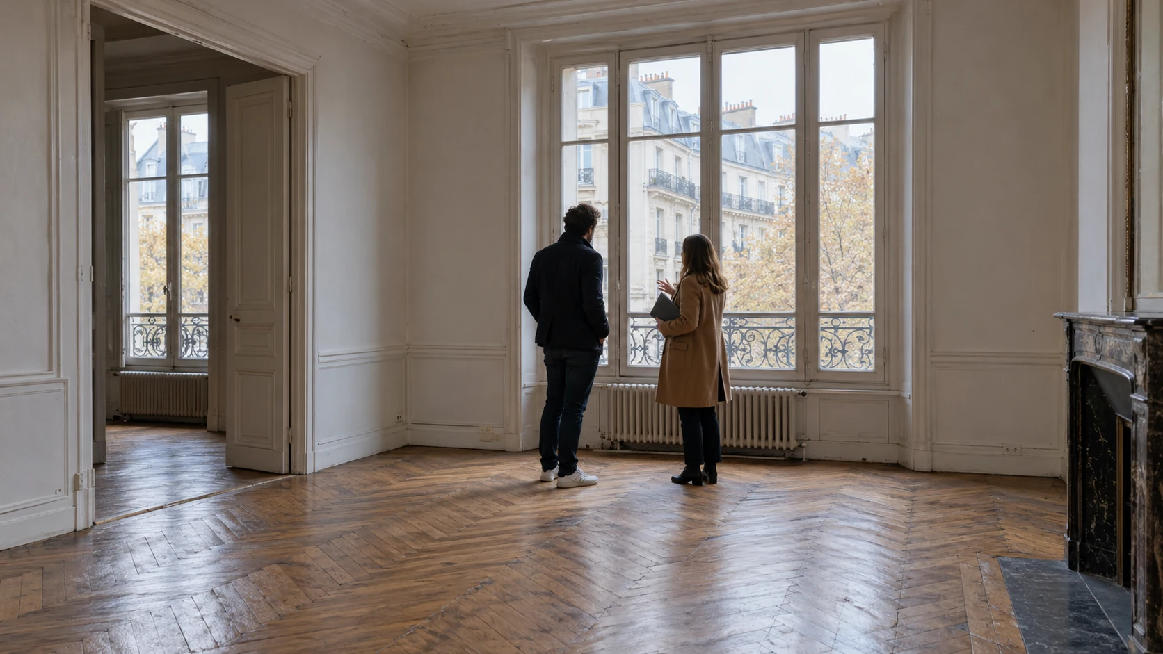 Deux personnes visitent un appartement ancien lumineux, près d’une fenêtre donnant sur des immeubles français.